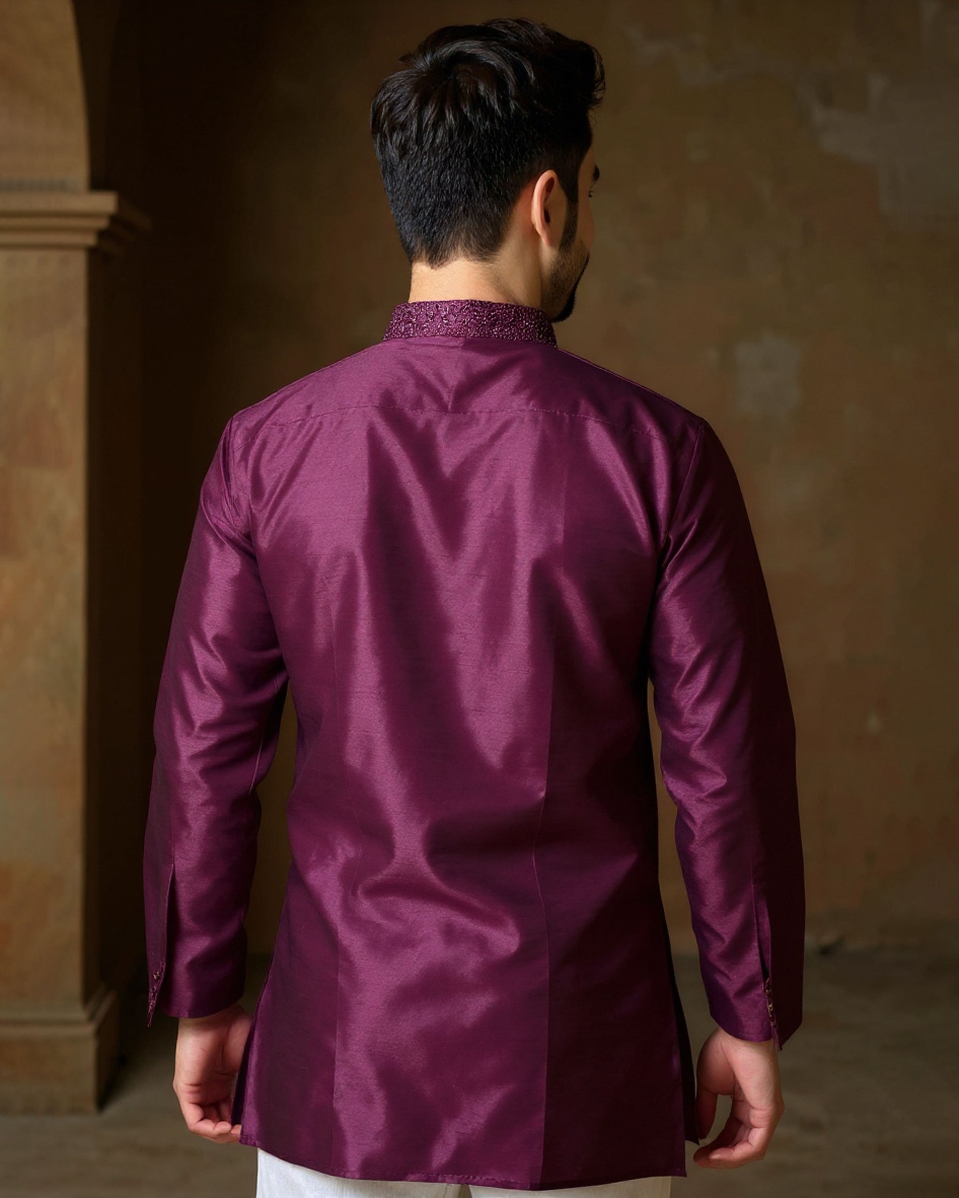 Man wearing a purple traditional outfit with a textured pattern, standing against a warm-toned wall.