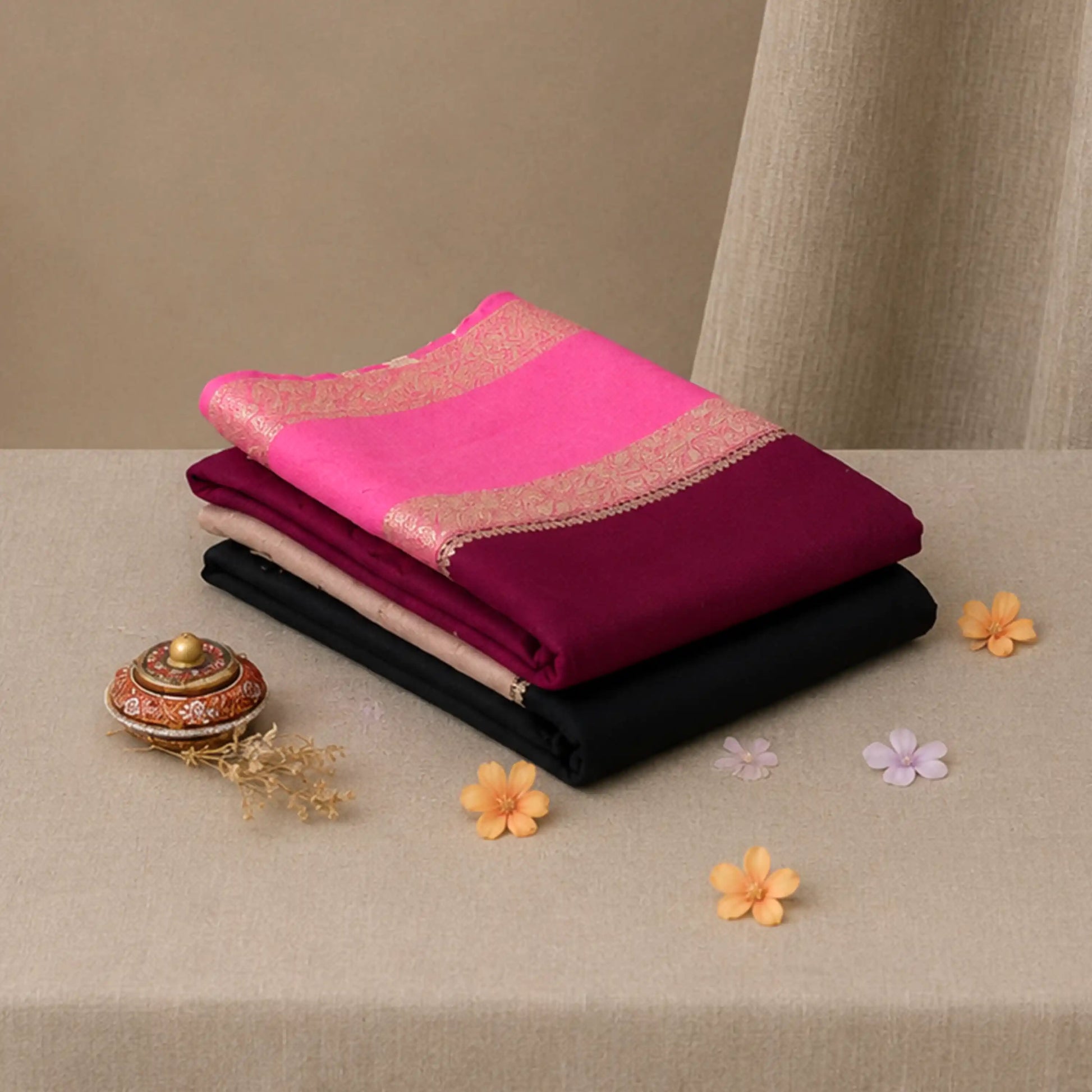 A stack of folded sarees in vibrant colors, featuring a bright pink saree with a gold border on top, followed by a  black saree. 