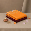 A stack of folded textiles, with the top item being a bright orange saree and the bottom item a deep maroon saree 
