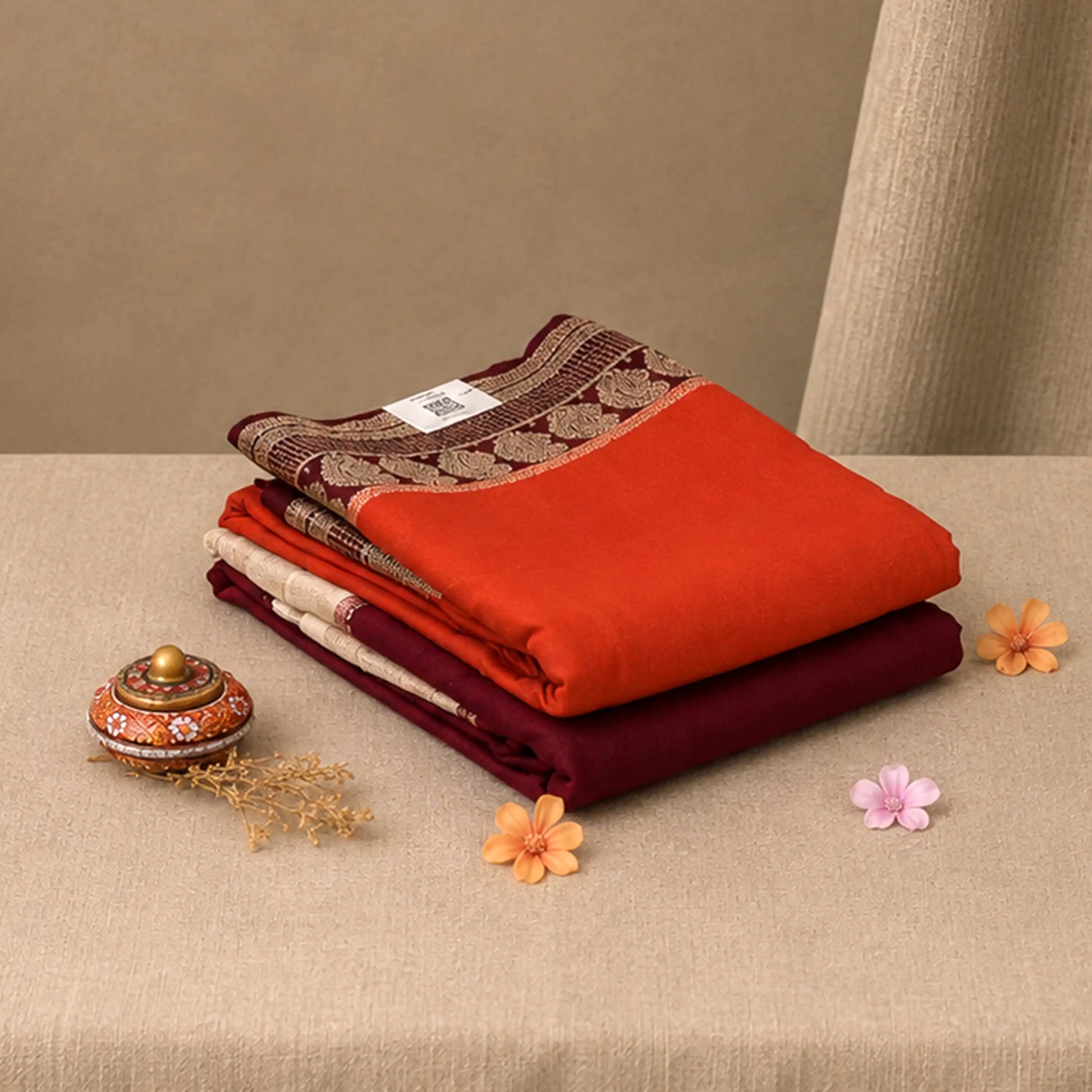 A stack of folded fabric items in warm tones, including a bright orange-red piece, deep burgundy, cream, and a brown patterned border