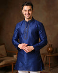 Man wearing a blue kurta with intricate designs in a warm-toned room.