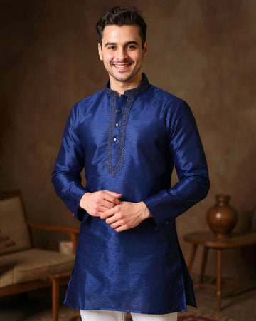 Man wearing a blue kurta with intricate designs in a warm-toned room.