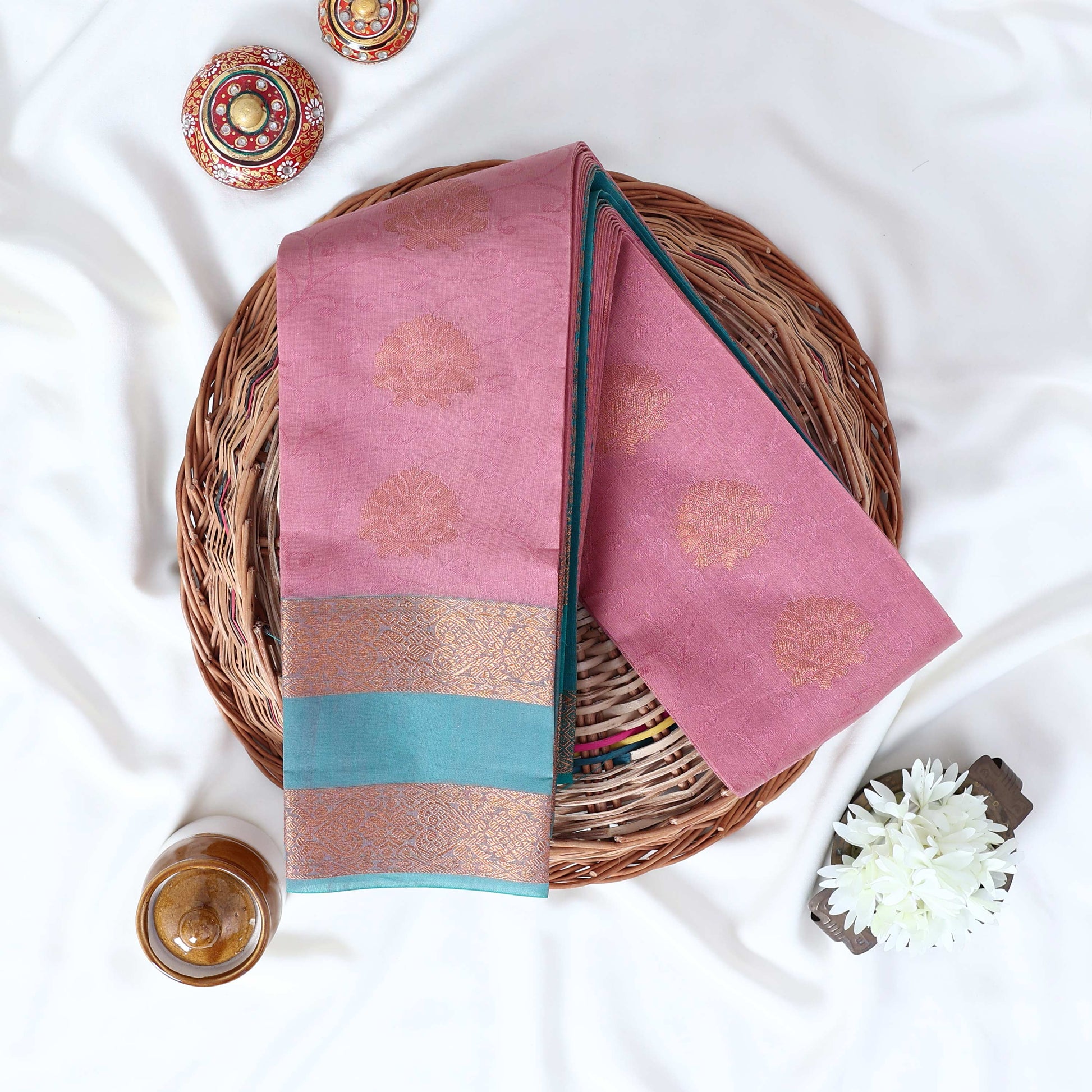 Elampillai silk cotton saree with floral butta motifs pink body and light blue zari border folded top view on woven basket.