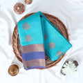 Elampillai silk cotton saree with floral butta motifs teal blue body and lavender zari border folded top view on woven basket.