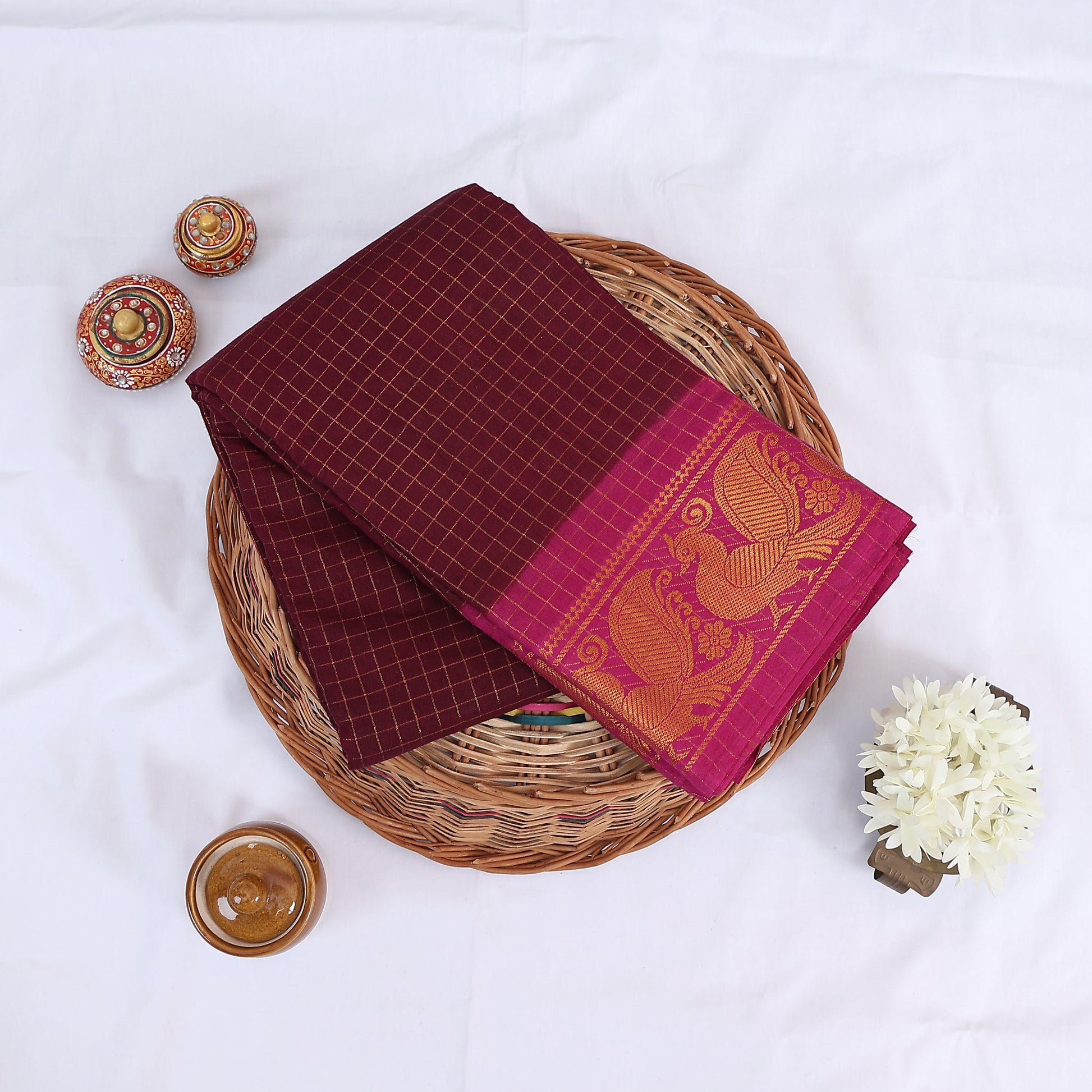 Sungudi cotton saree with gold zari checks body and maroon peacock motif border folded top view on wicker basket