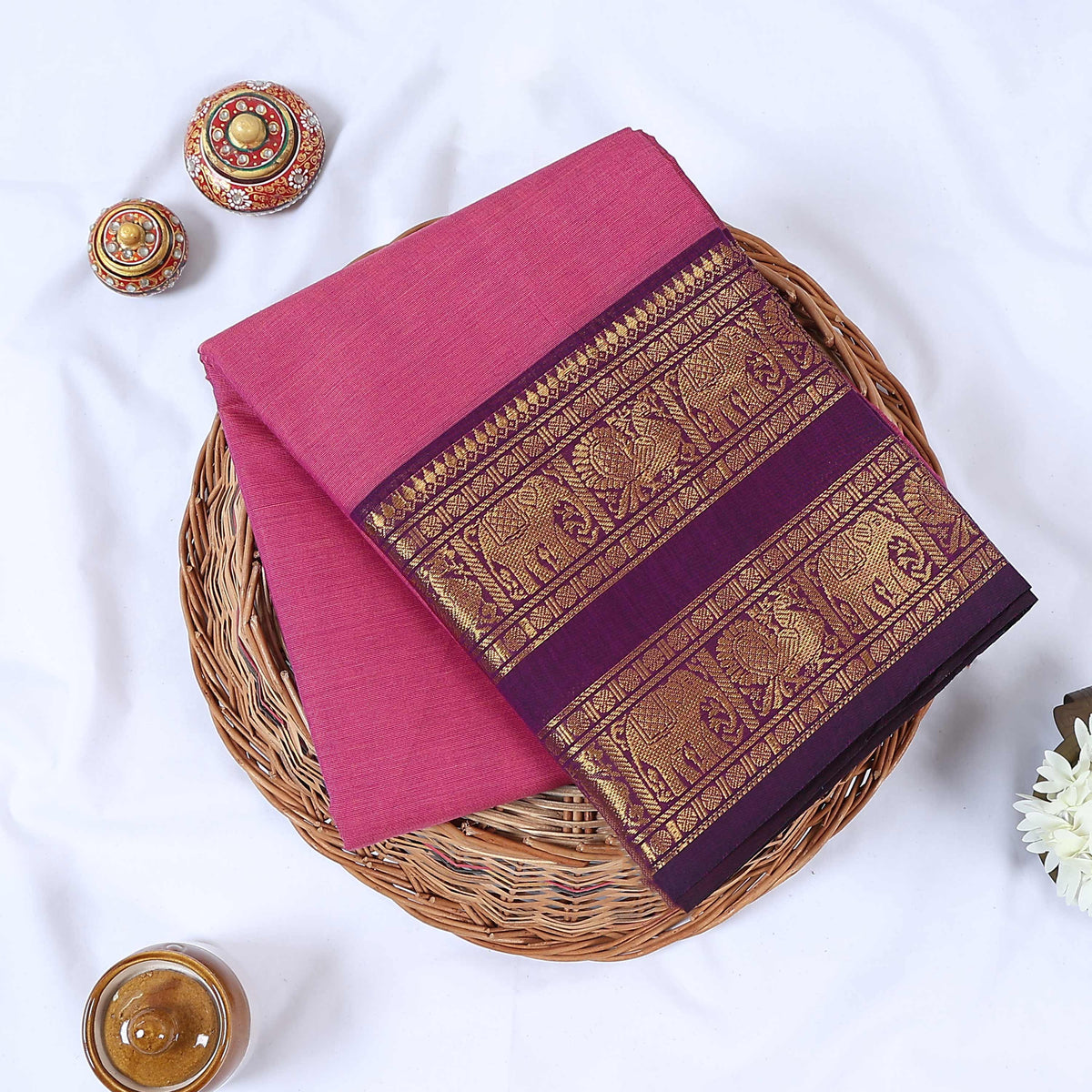 Venkatagiri cotton saree with purple zari border in pink folded flat on a wicker basket top view