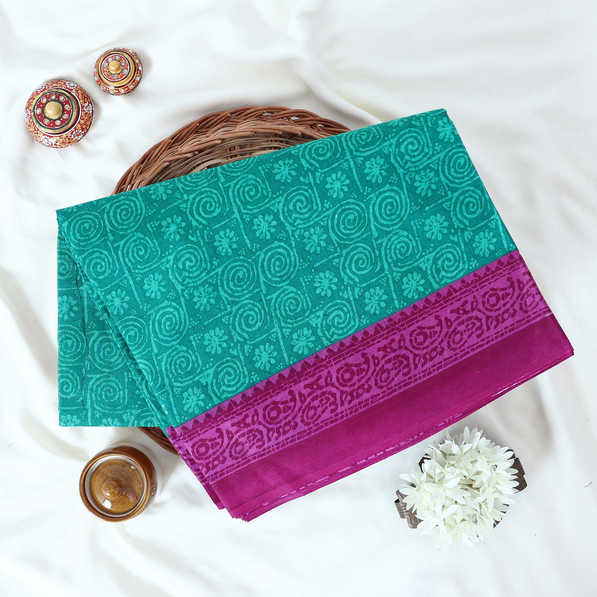 Green Voile cotton saree with spiral and floral printed motifs contrast border front flat lay view on a woven basket.