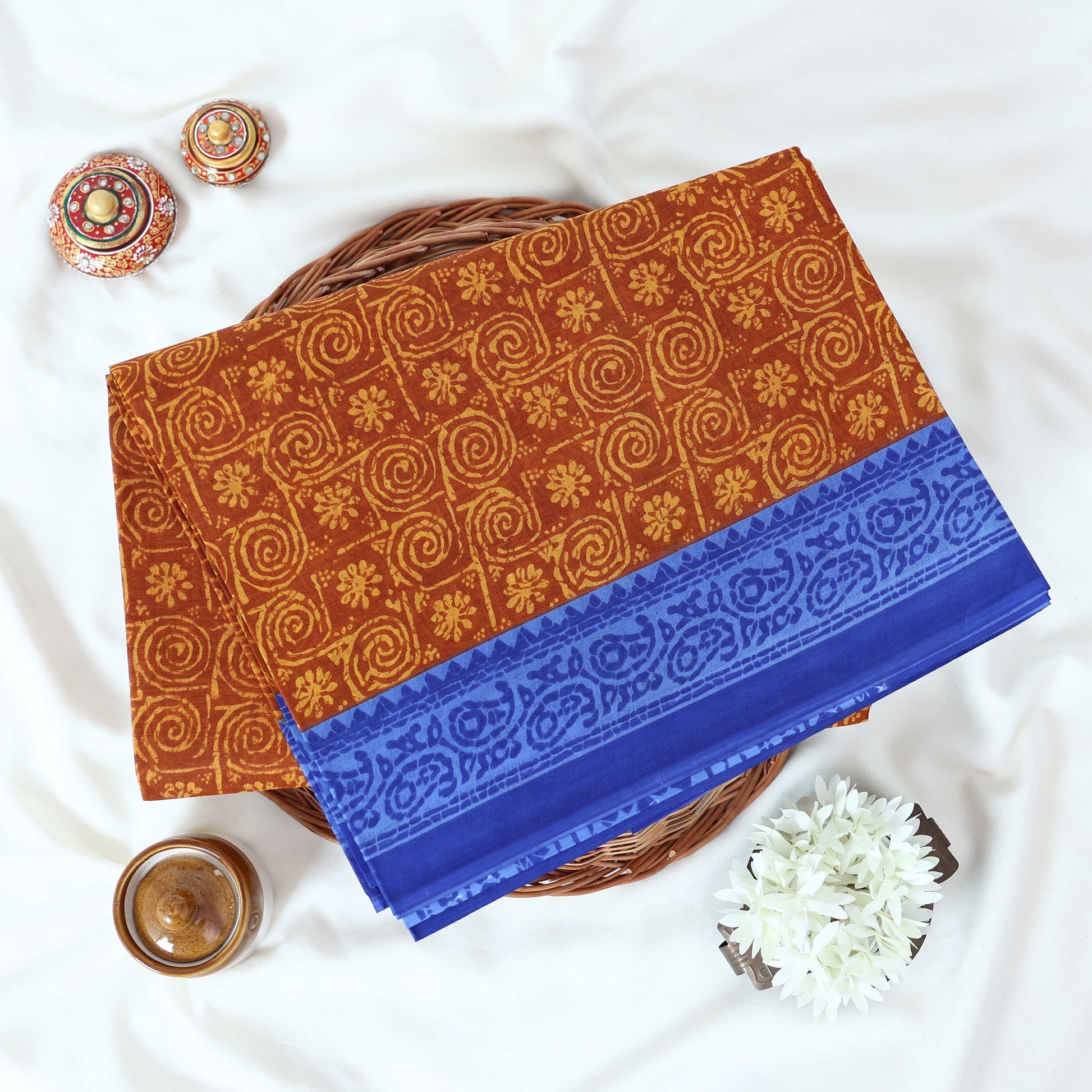 Brown Voile cotton saree with spiral and floral printed motifs contrast border front flat lay view on a woven basket.