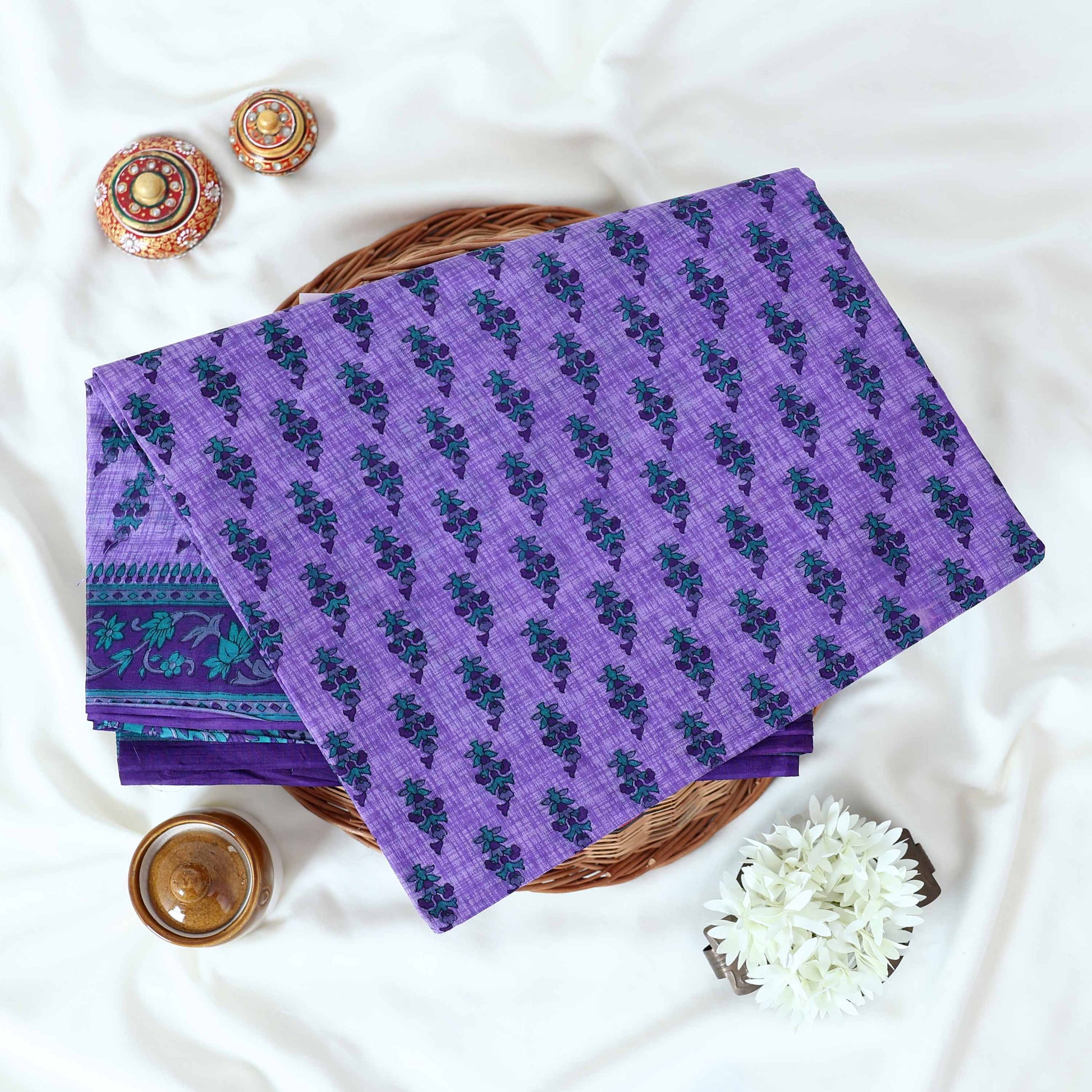 Voile cotton saree with floral motif print and decorative border in purple folded flat lay view on woven basket.