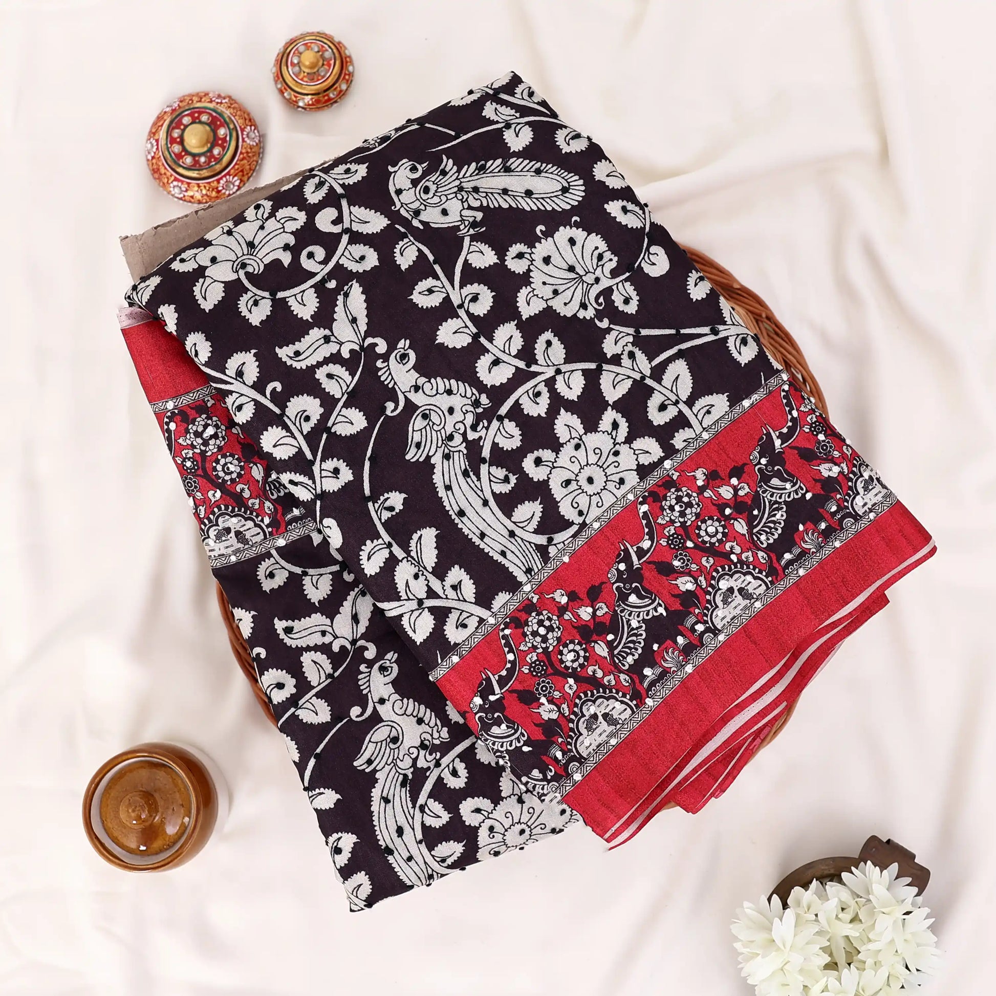 A folded black and red printed saree features a red base with black and white floral patterns and a solid red border.