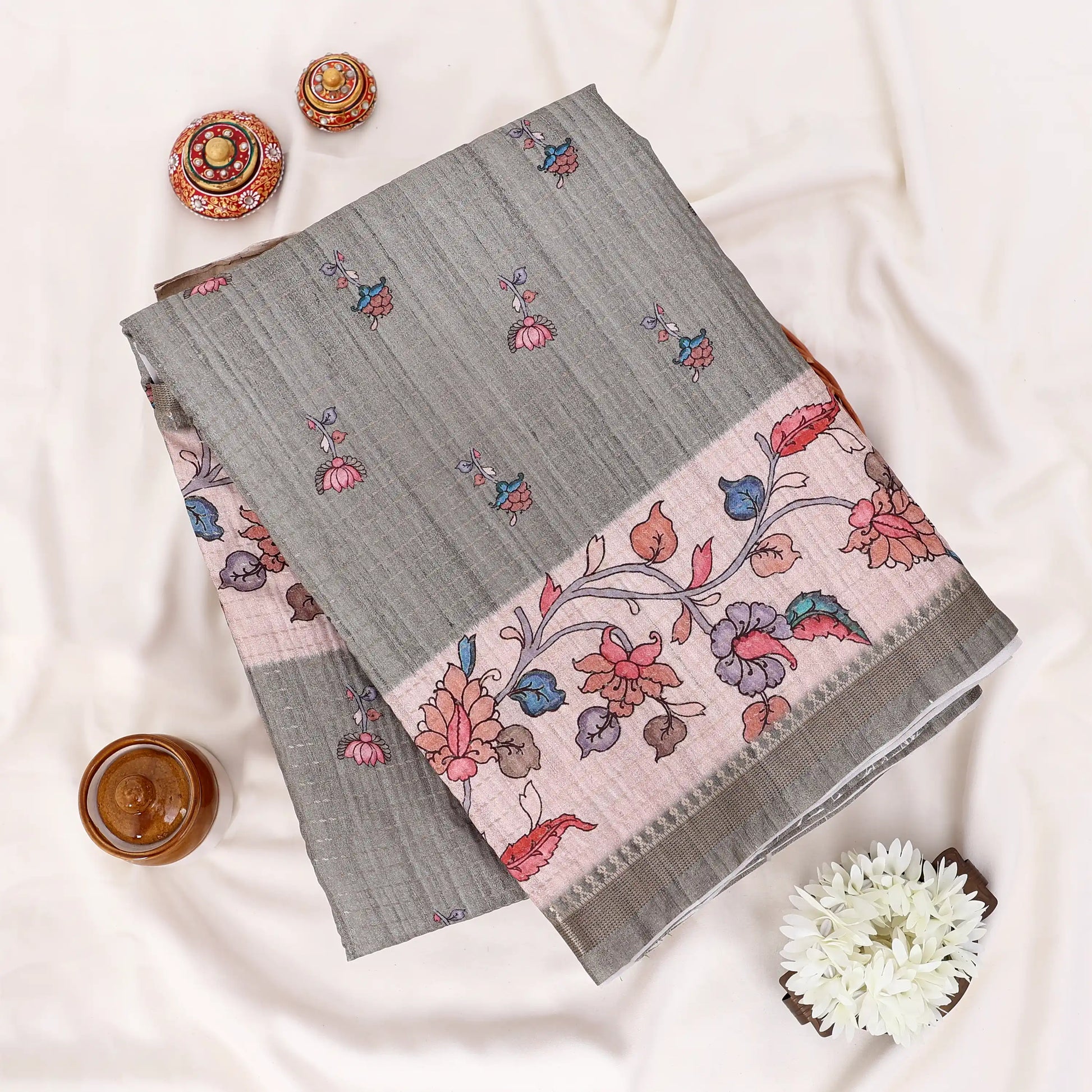 A folded gray saree with a pink border featuring colorful floral and bird motifs