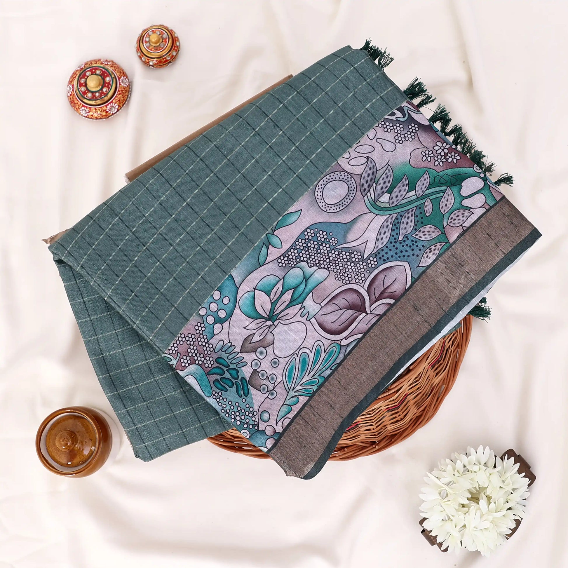 A folded patterned saree item, with teal-green grid pattern on one section, a central panel with a floral and paisley print 