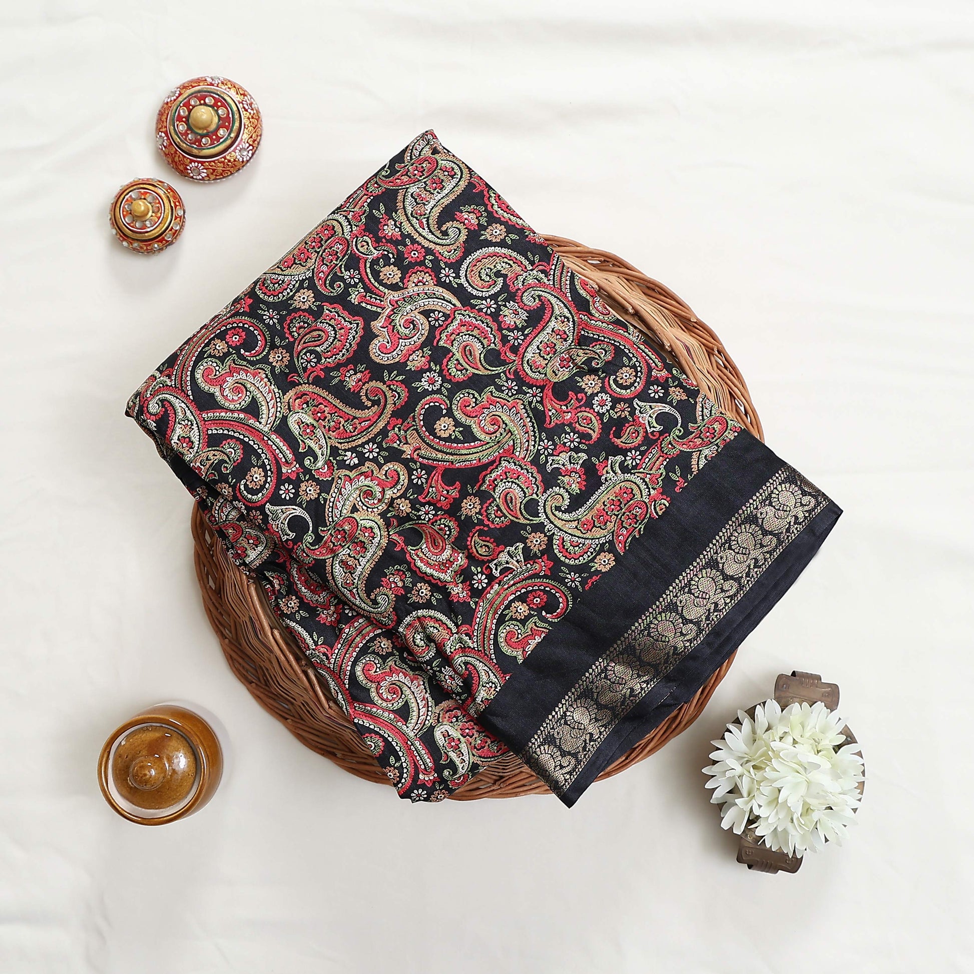 Art silk saree with intricate paisley floral print in black and gold woven border folded flat lay view in a wicker basket
