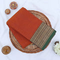 Chettinad cotton saree with rust orange body and green zari border folded top view with woven texture.