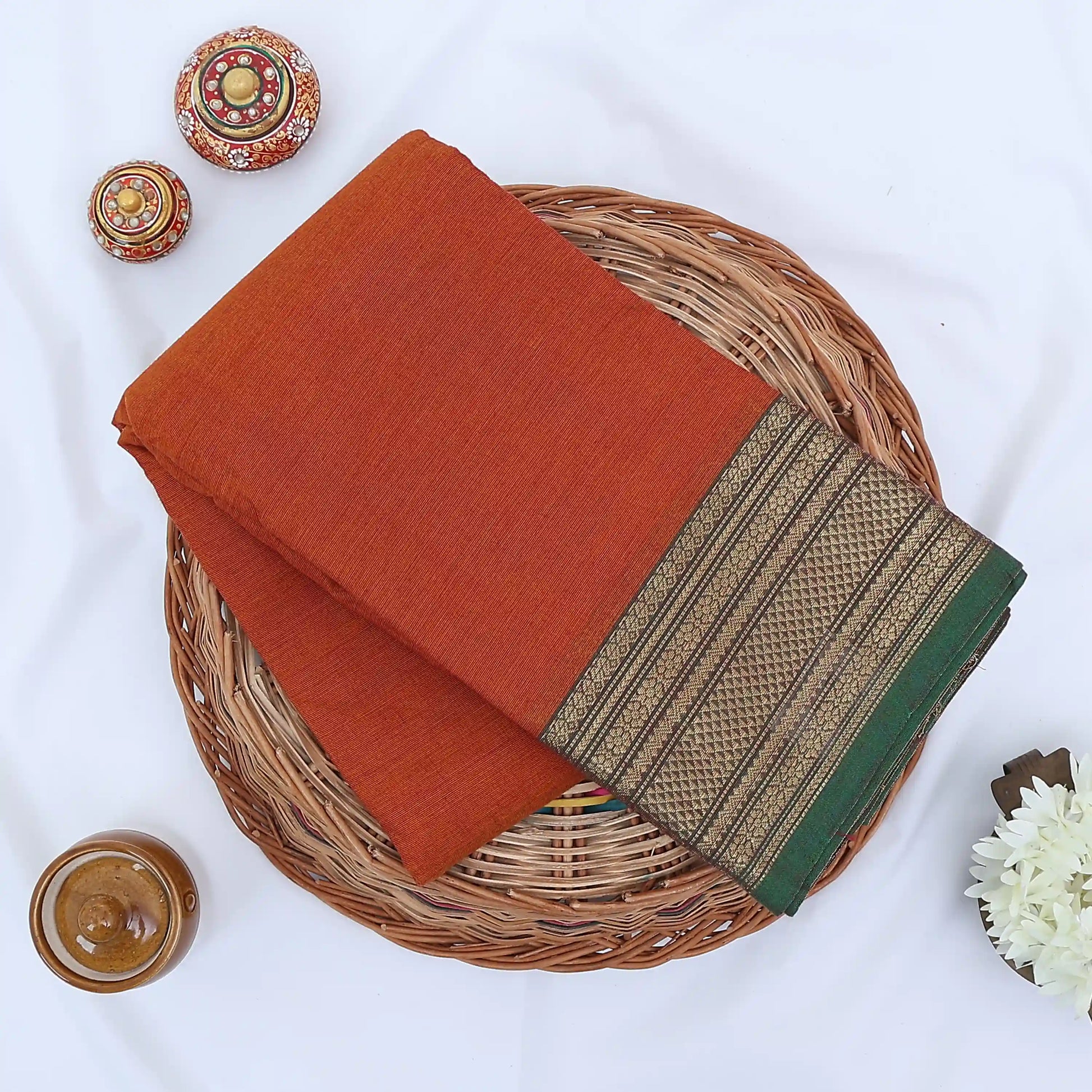 Chettinad cotton saree with rust orange body and green zari border folded top view with woven texture.