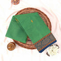 Kanchi Cotton Saree In Green With Blue And Gold Woven Border Neatly Folded On Wicker Basket Top View Product Display
