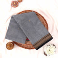 Kanchi Cotton Saree With Grey And Black Gold Zari Border Folded Flat Lay View On Wicker Tray With Decor Accents
