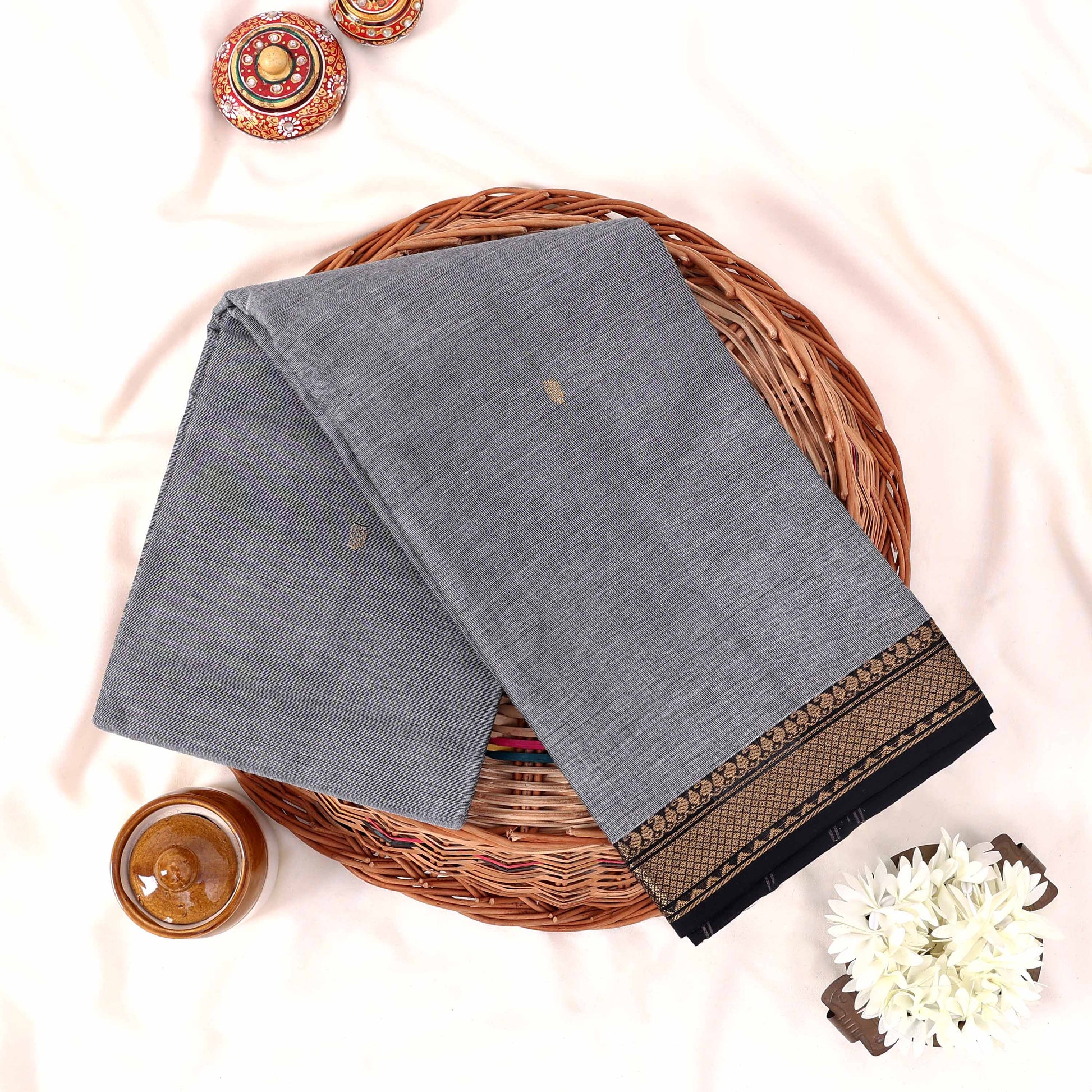 Kanchi Cotton Saree With Grey And Black Gold Zari Border Folded Flat Lay View On Wicker Tray With Decor Accents
