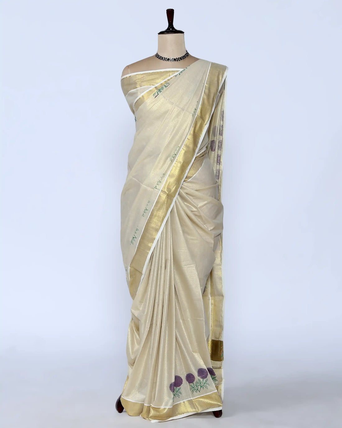 A cream-colored kerala saree with a metallic gold border and purple floral embroidery near the hem full view