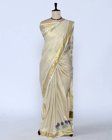 A cream-colored kerala saree with a metallic gold border and purple floral embroidery near the hem full view