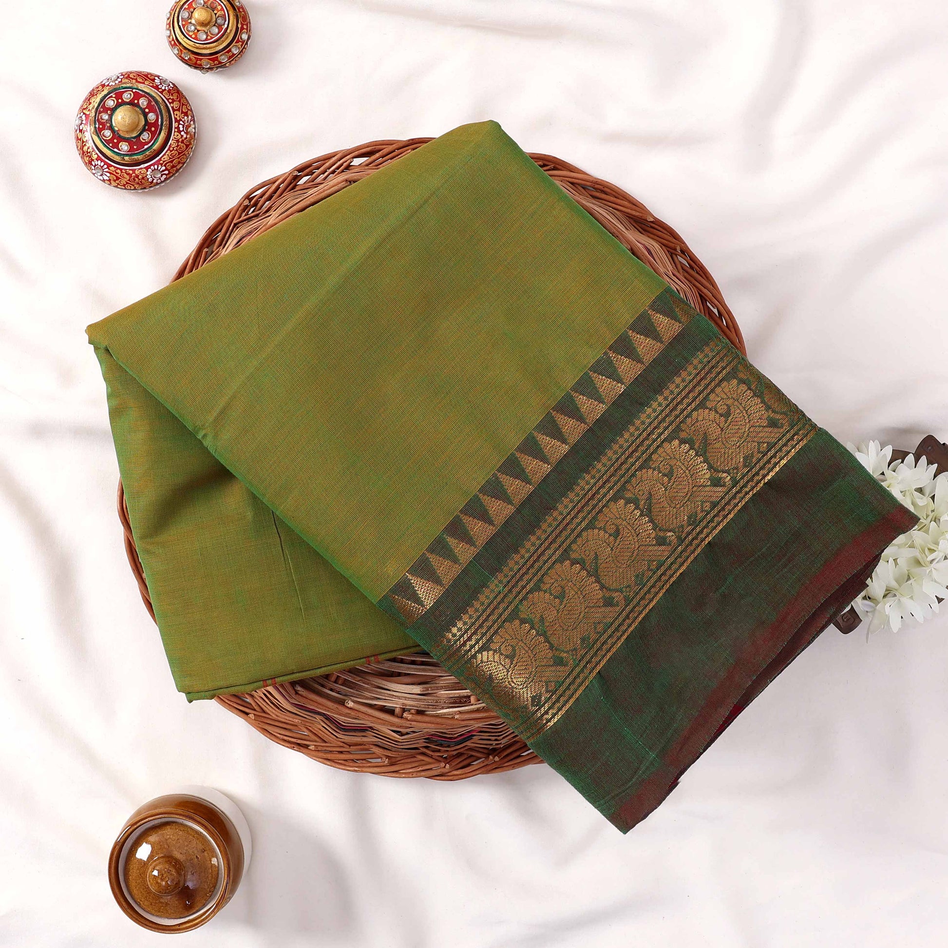 Kanchi Cotton Saree With Light Green Body And Ornate Gold Woven Border Folded On A Wicker Tray Top View Display
