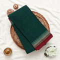 Narayanpet Cotton saree with plain deep green body and maroon zari border folded close up view on a woven basket display.