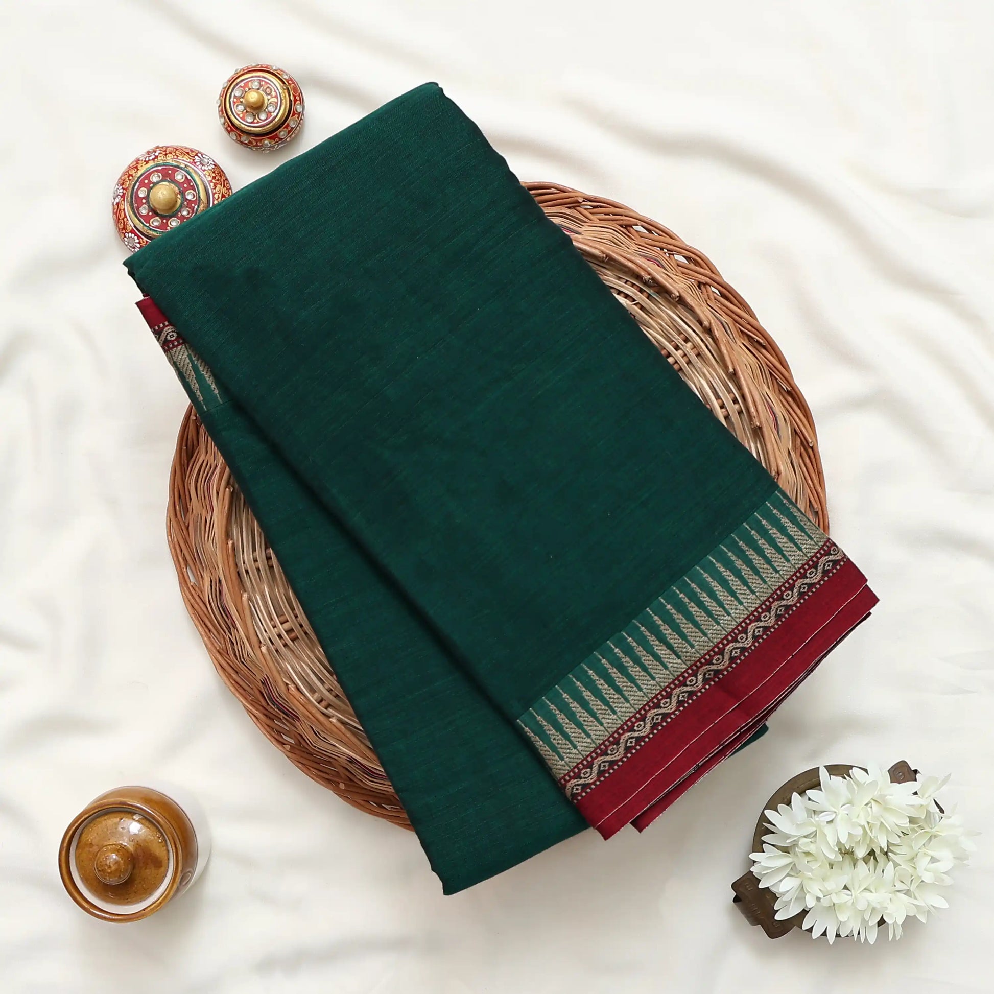 Narayanpet Cotton saree with plain deep green body and maroon zari border folded close up view on a woven basket display.