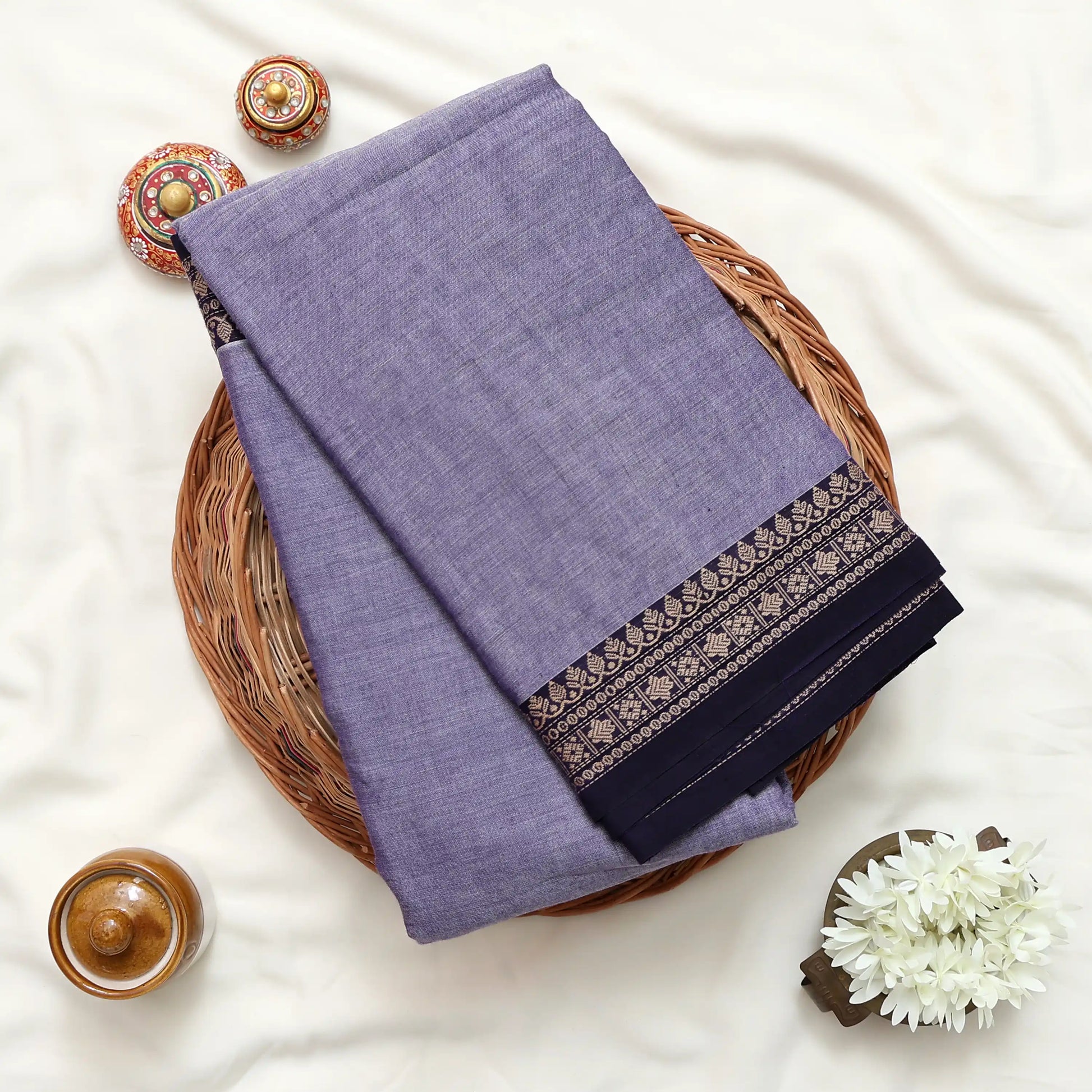Top view of lavender Narayanpet cotton saree with contrast zari woven border neatly folded on a wicker basket display.