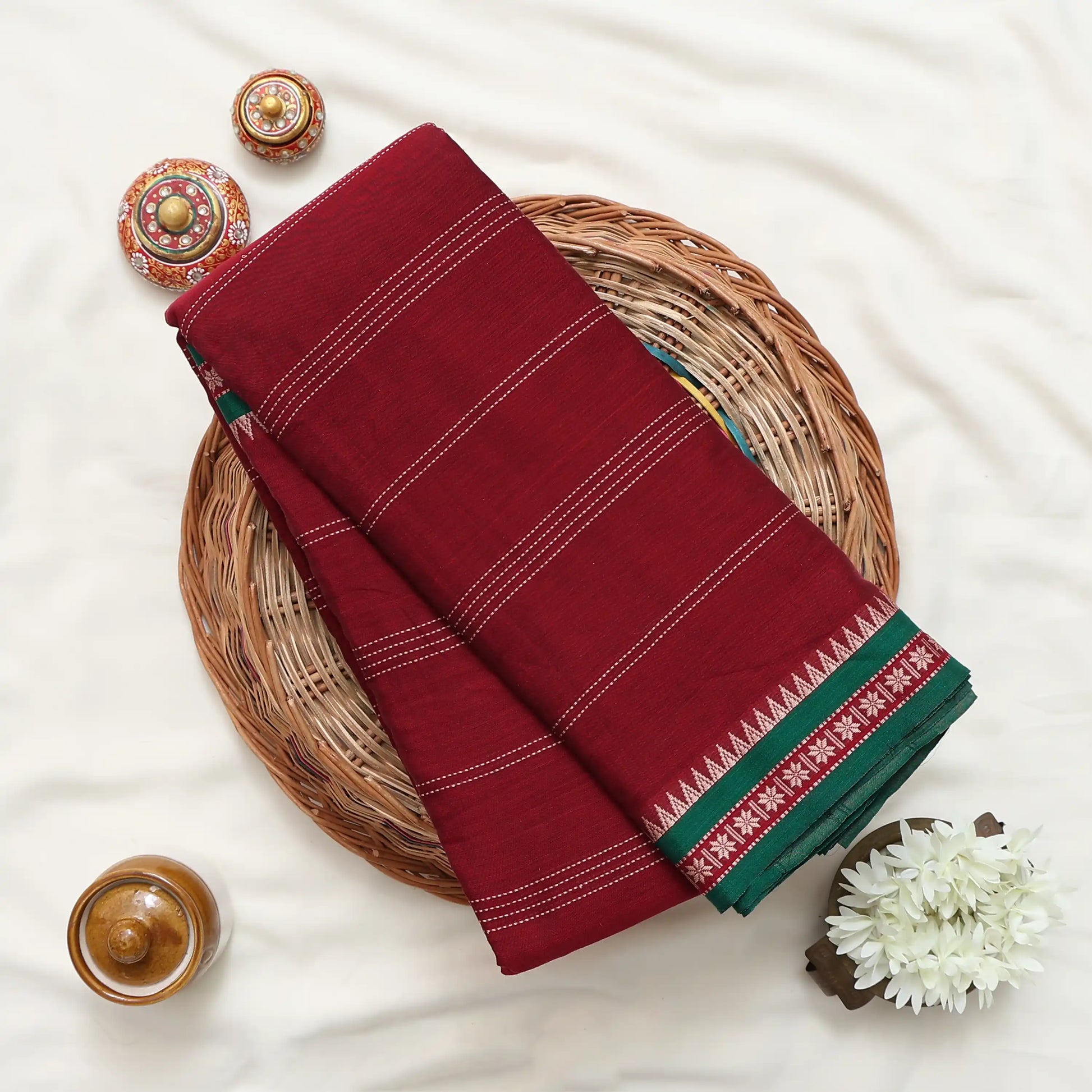A folded deep red saree  with white stripe details and a green patterned border