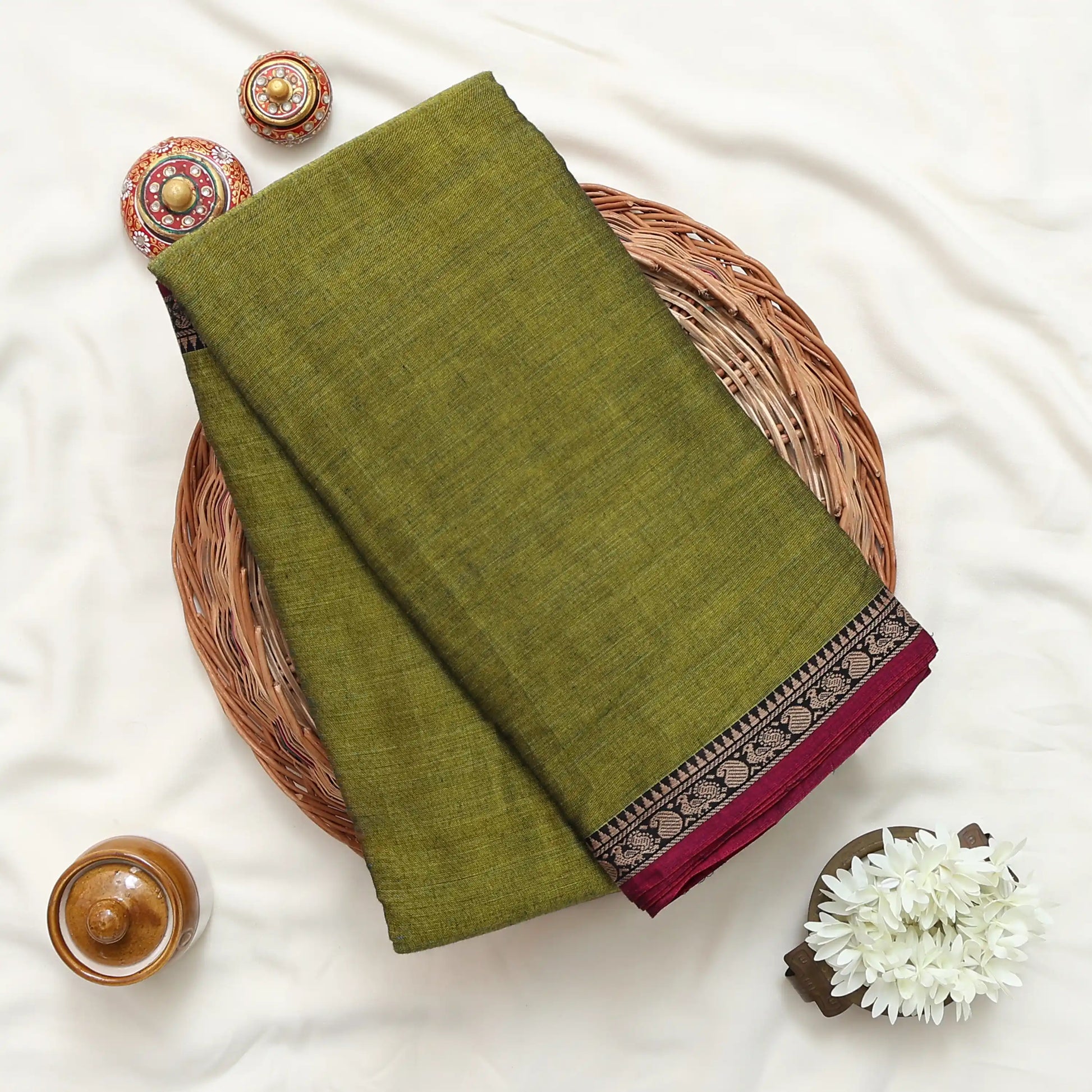 Narayanpet cotton saree with plain olive green body and paisley zari woven border folded top view on wicker basket.
