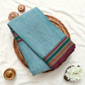 Top view of teal Narayanpet cotton saree with contrast striped zari woven border neatly folded on a wicker basket display.