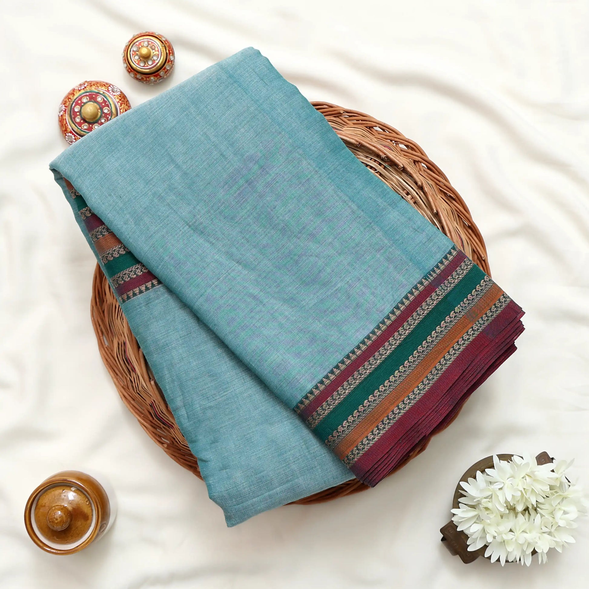 Top view of teal Narayanpet cotton saree with contrast striped zari woven border neatly folded on a wicker basket display.
