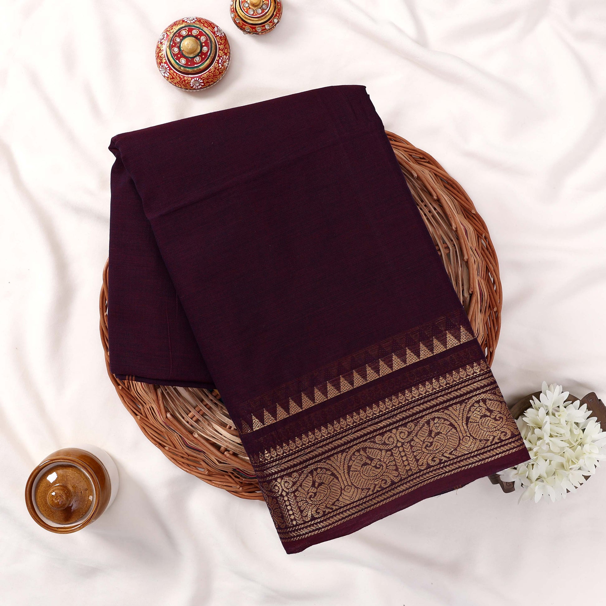 Kanchi Cotton Saree With Intricate Gold Zari Border In Deep Purple Neatly Folded On Wicker Basket Top View
