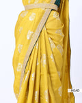 A close-up of a yellow saree with white floral motifs and a metallic gold border, draped elegantly. 