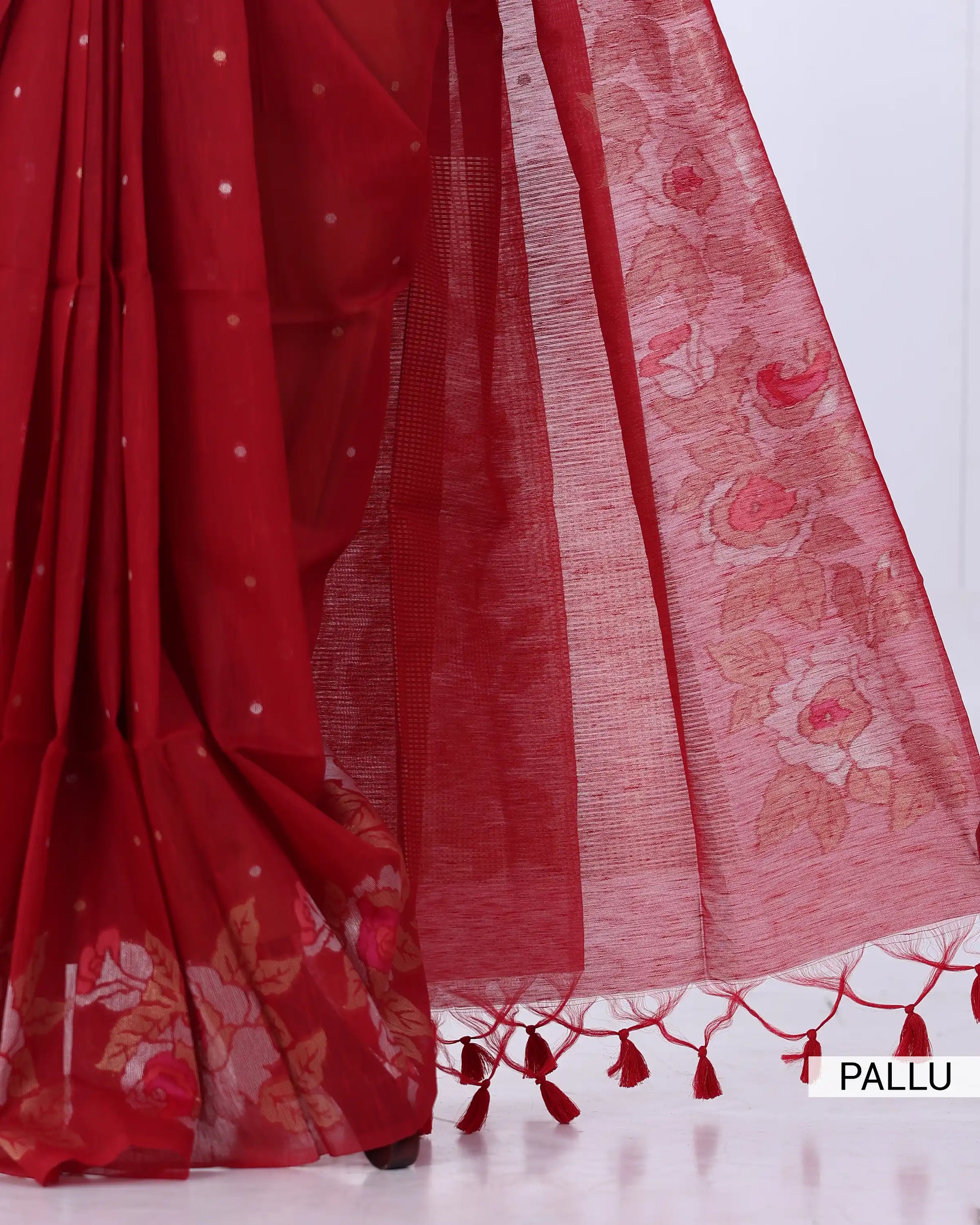 A close-up of a red saree with a matching pallu, showing floral printed detailing, sheer texture, and red tassels along the edge