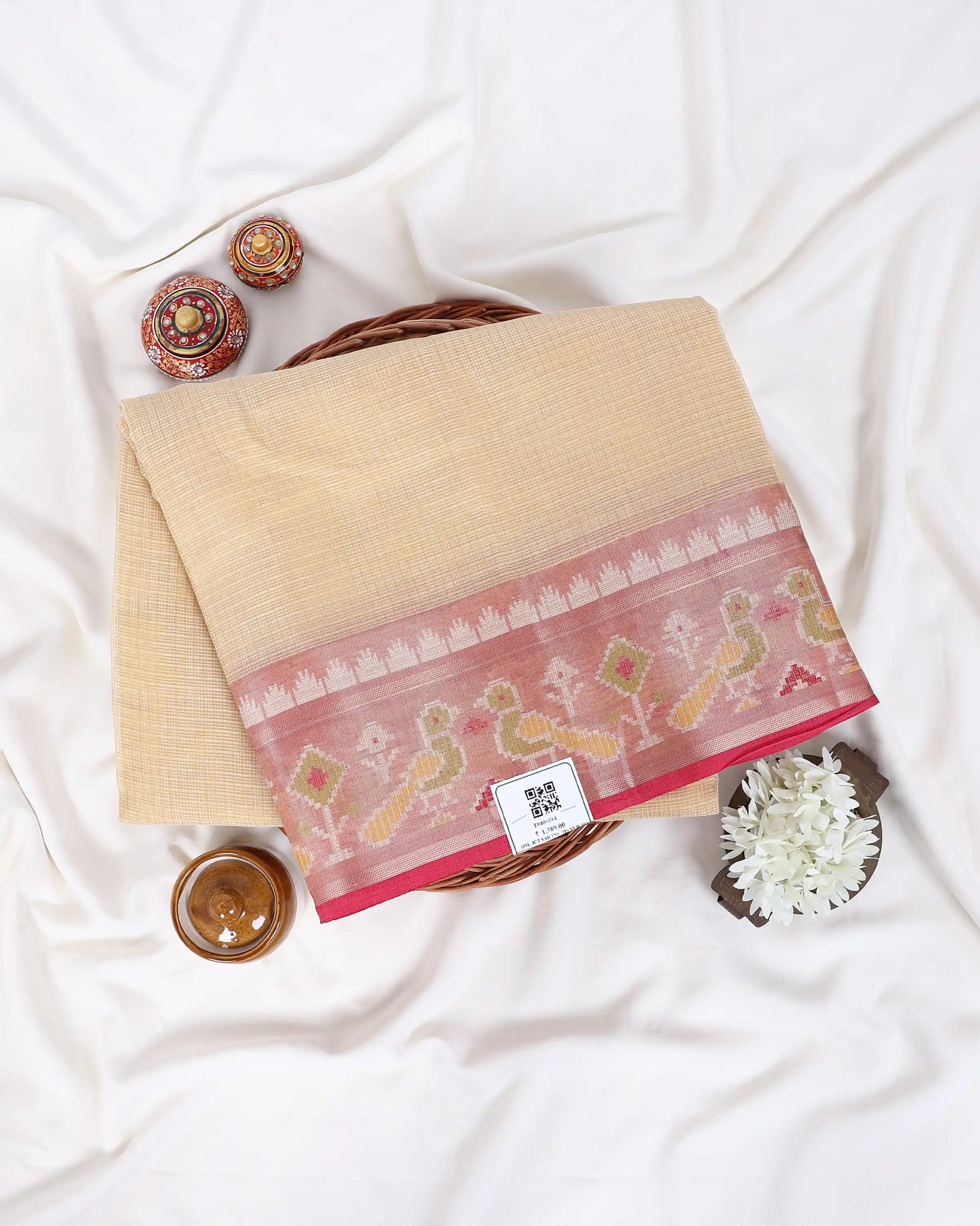 A folded beige cotton saree with a pink border featuring traditional bird and floral motifs