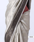 A close-up of a draped saree in light beige with a wide dark gray border featuring intricate paisley and floral zari patterns, with a hint of red fabric visible at the edge.