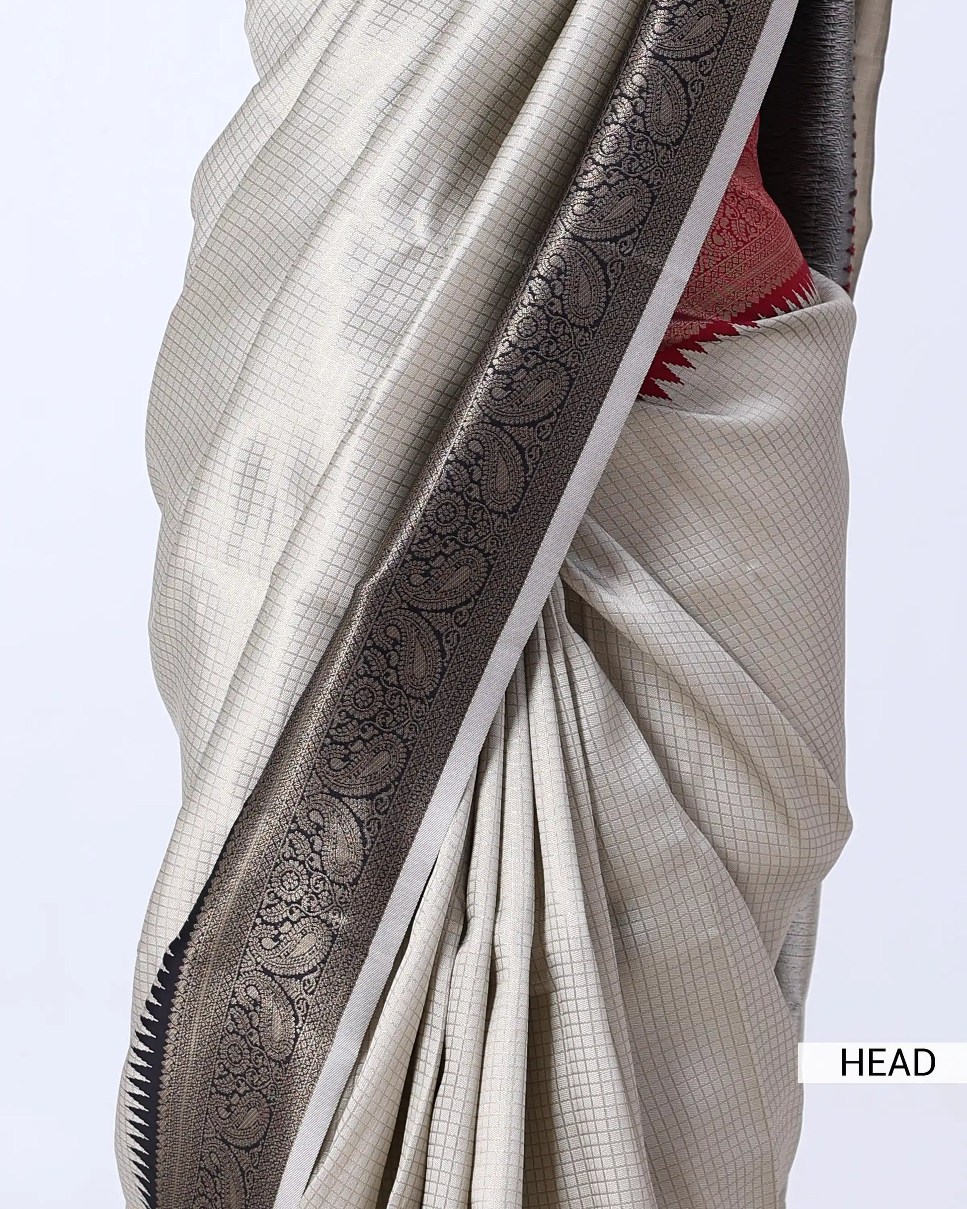 A close-up of a draped saree in light beige with a wide dark gray border featuring intricate paisley and floral zari patterns, with a hint of red fabric visible at the edge.