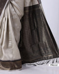 A close-up of a traditional Indian saree, showing the off-white body fabric, the dark brown patterned pallu border, and the black tassel fringe.