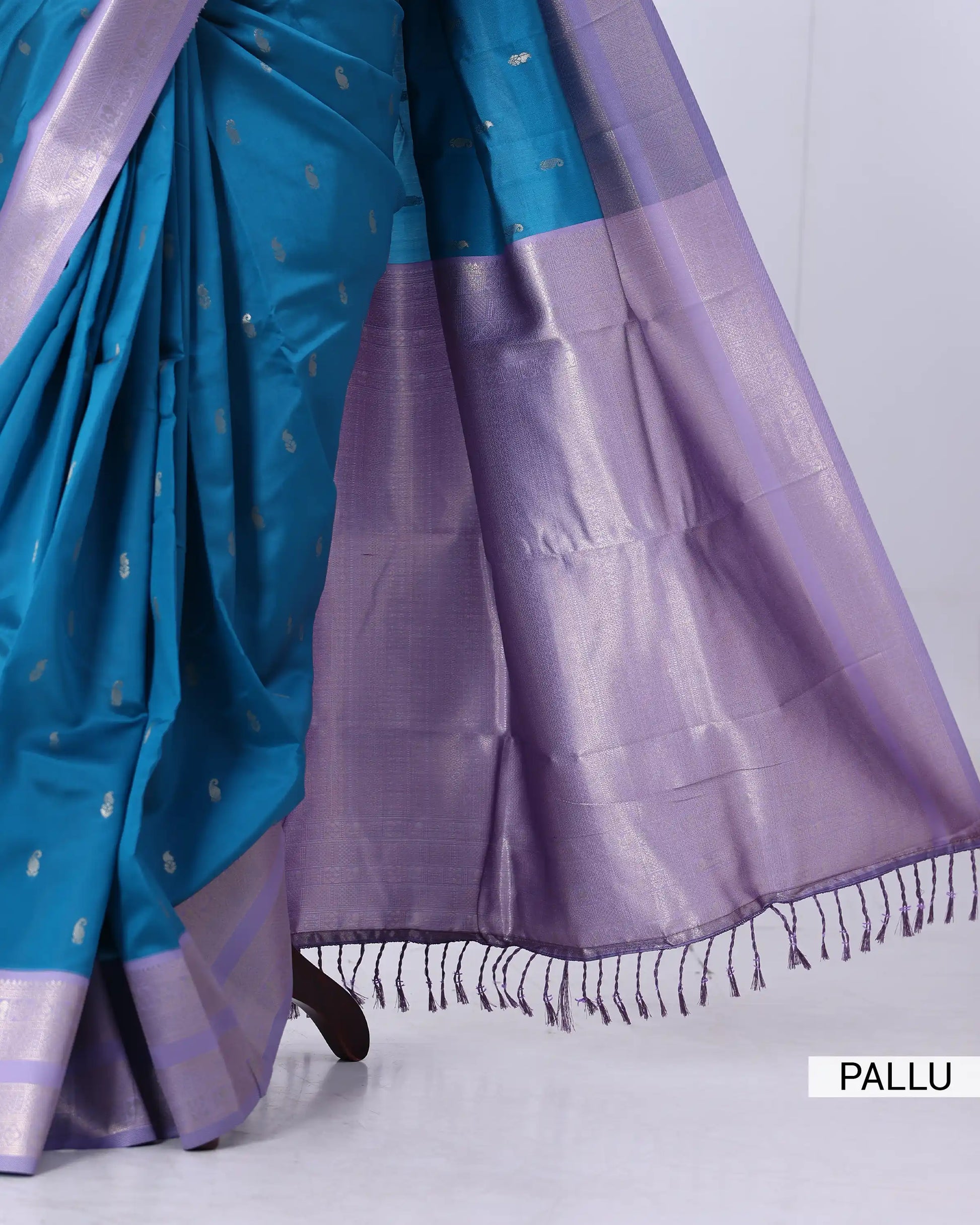 A close-up of a draped saree with a teal blue body, small metallic motifs, and a wide lavender-purple pallu border with fringe