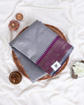 A folded gray silk saree with a purple and gold border is displayed on a round woven tray 