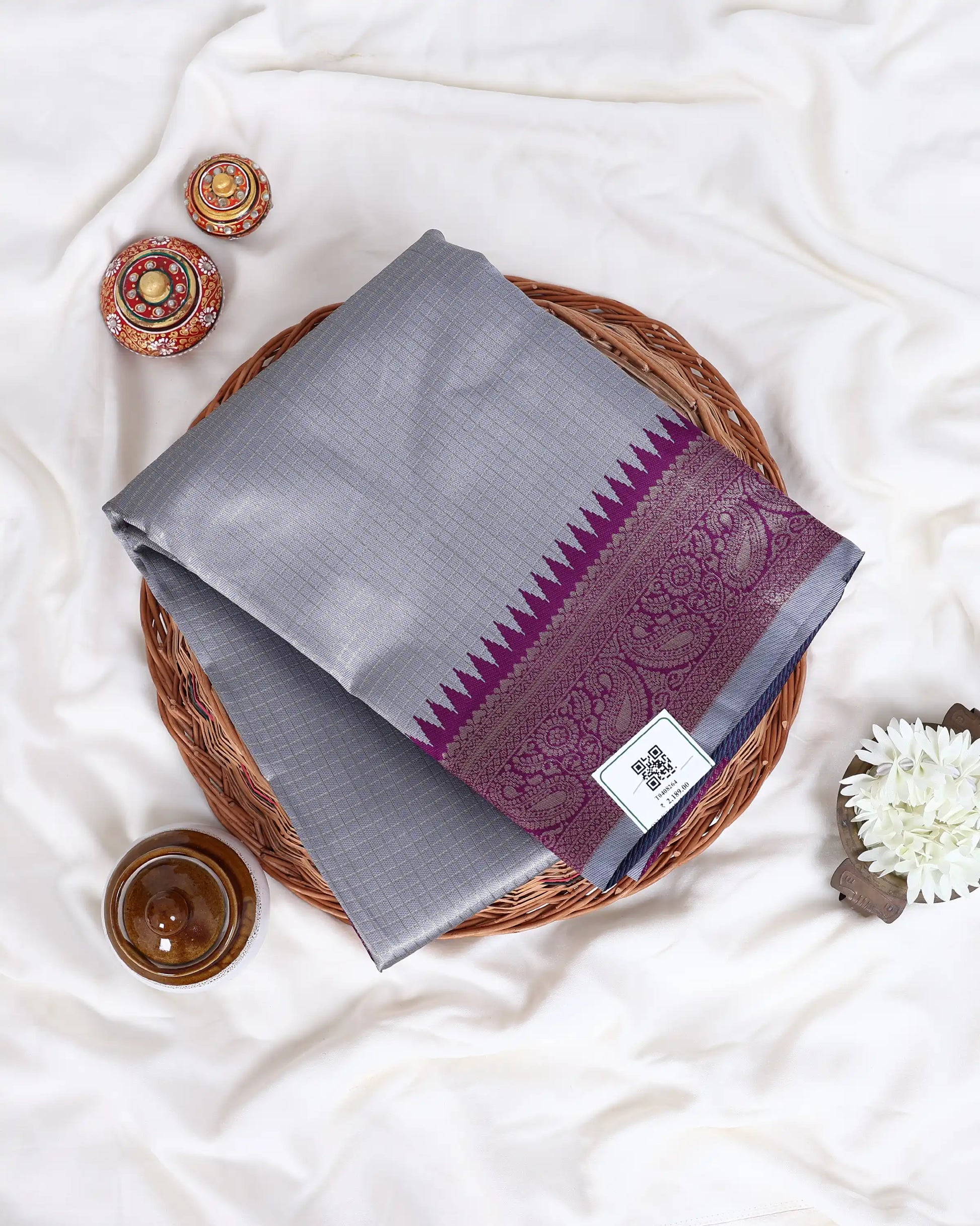A folded gray silk saree with a purple and gold border is displayed on a round woven tray 