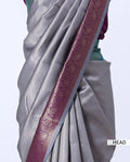 A close-up of a draped saree in light silver-gray with a wide metallic magenta border featuring gold paisley motifs