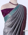 A grey silk saree draped on a mannequin, featuring a deep purple blouse with a teal and gold border, and a matching purple border on the saree.