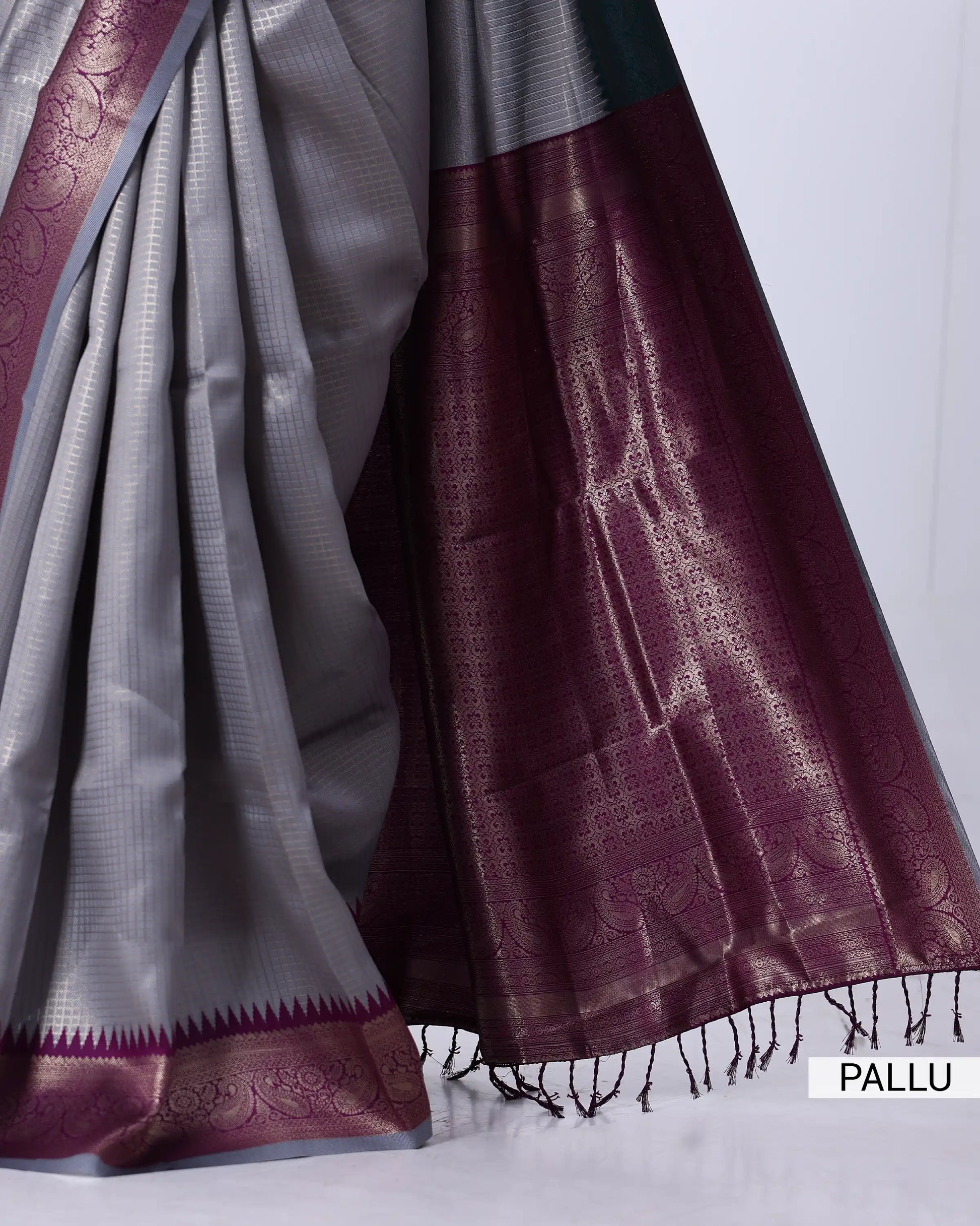 A close-up of a traditional Indian saree, showing the silver-gray body fabric, the deep maroon border, and the metallic gold zari woven patterns.