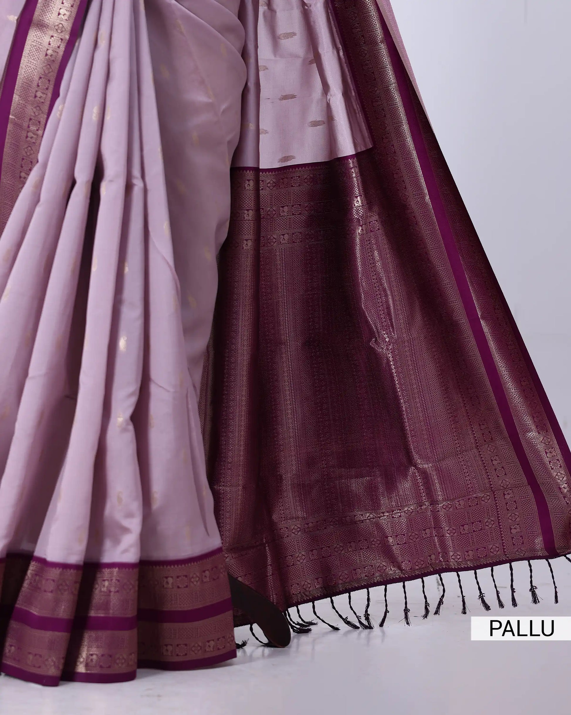 A close-up of a traditional Indian saree, showing the pallu end. The fabric is a light lavender-pink color with a rich, dark plum purple border and a metallic gold zari border.
