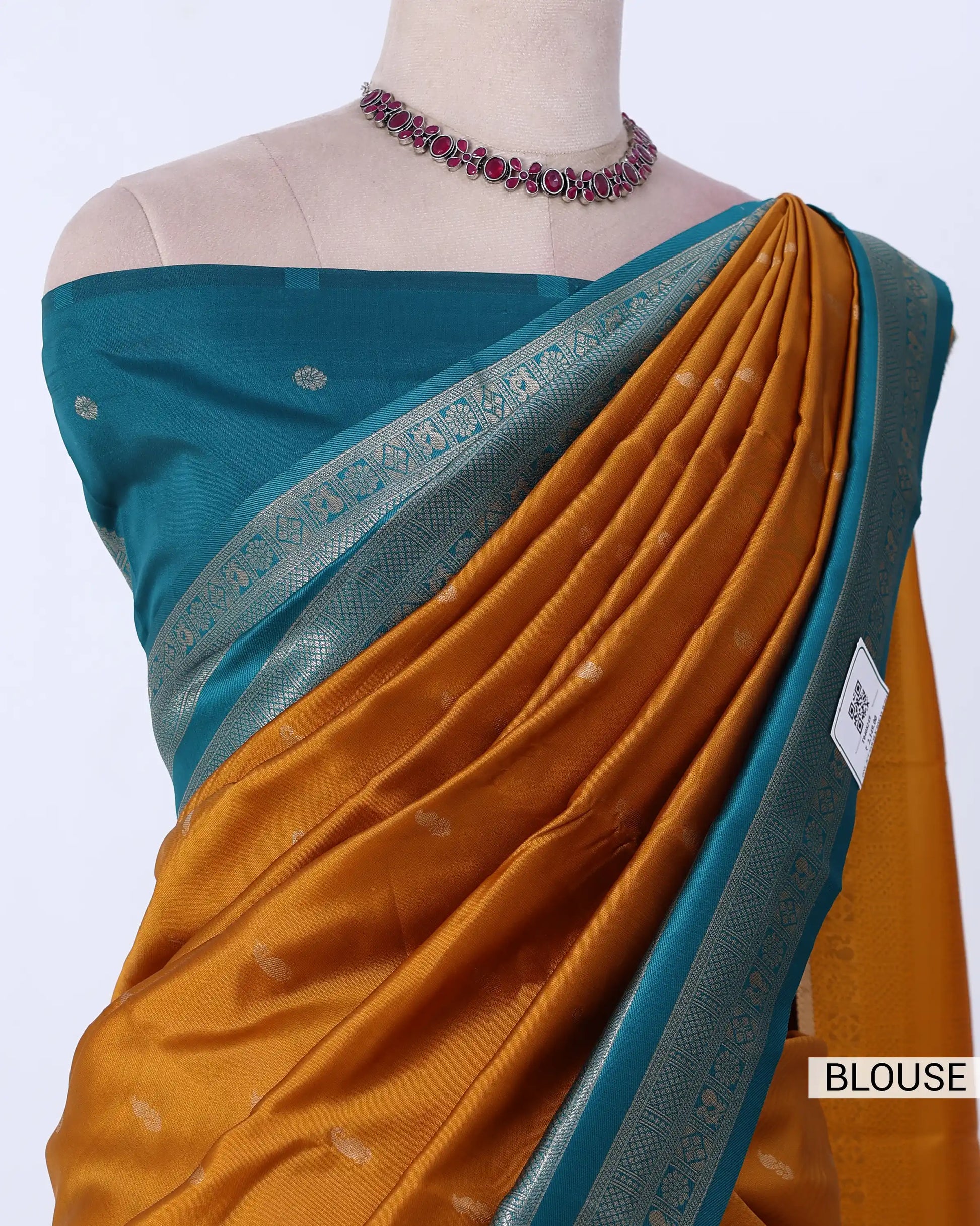 A close-up of a blouse and sari set displayed on a mannequin torso. The blouse is teal blue with small metallic motifs, paired with a mustard yellow-orange sari featuring a teal border and subtle woven dots.