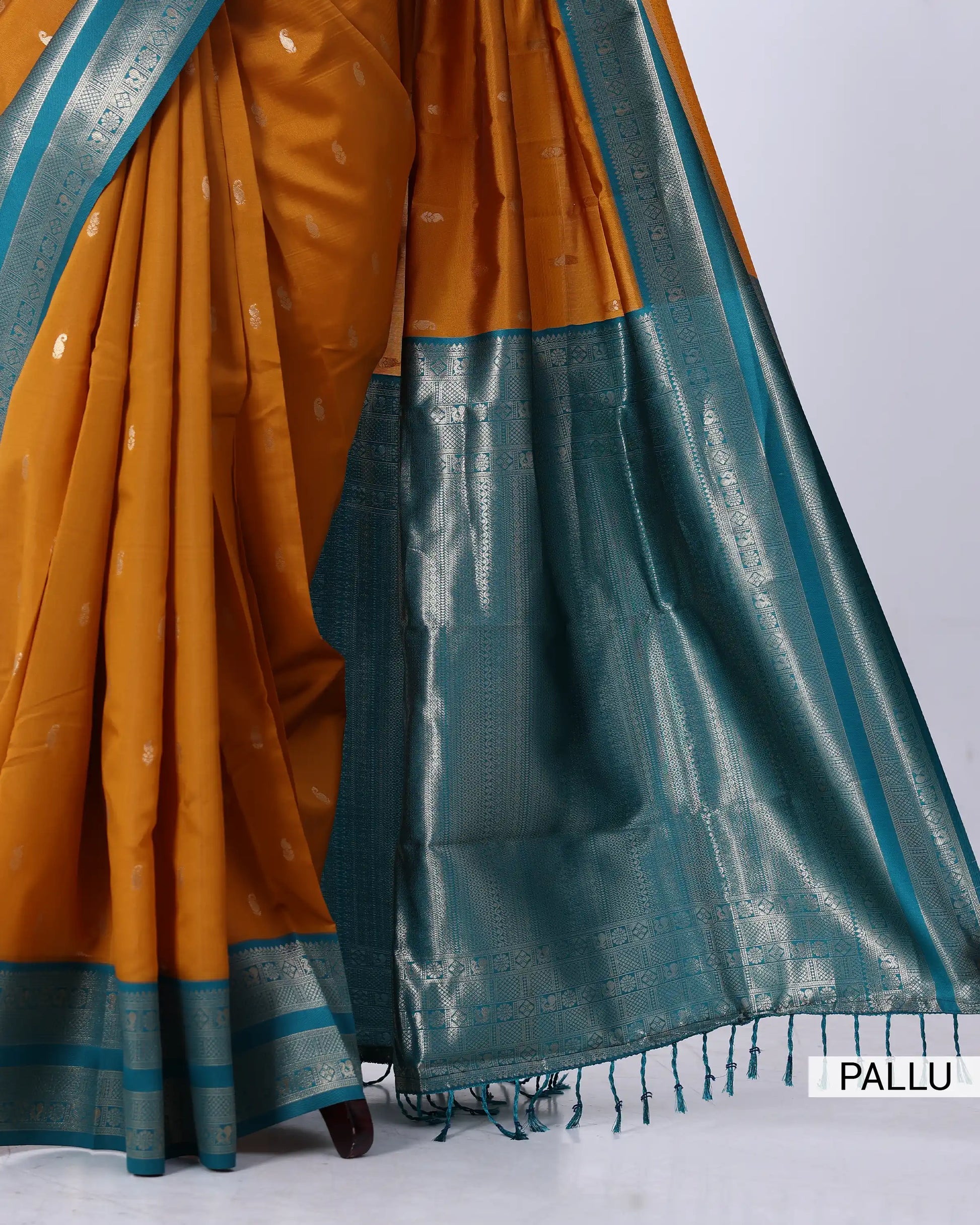 A close-up of a traditional Indian saree, showing its mustard yellow body with small metallic motifs, a teal blue border, and a wide teal blue pallu with silver zari weaving. 