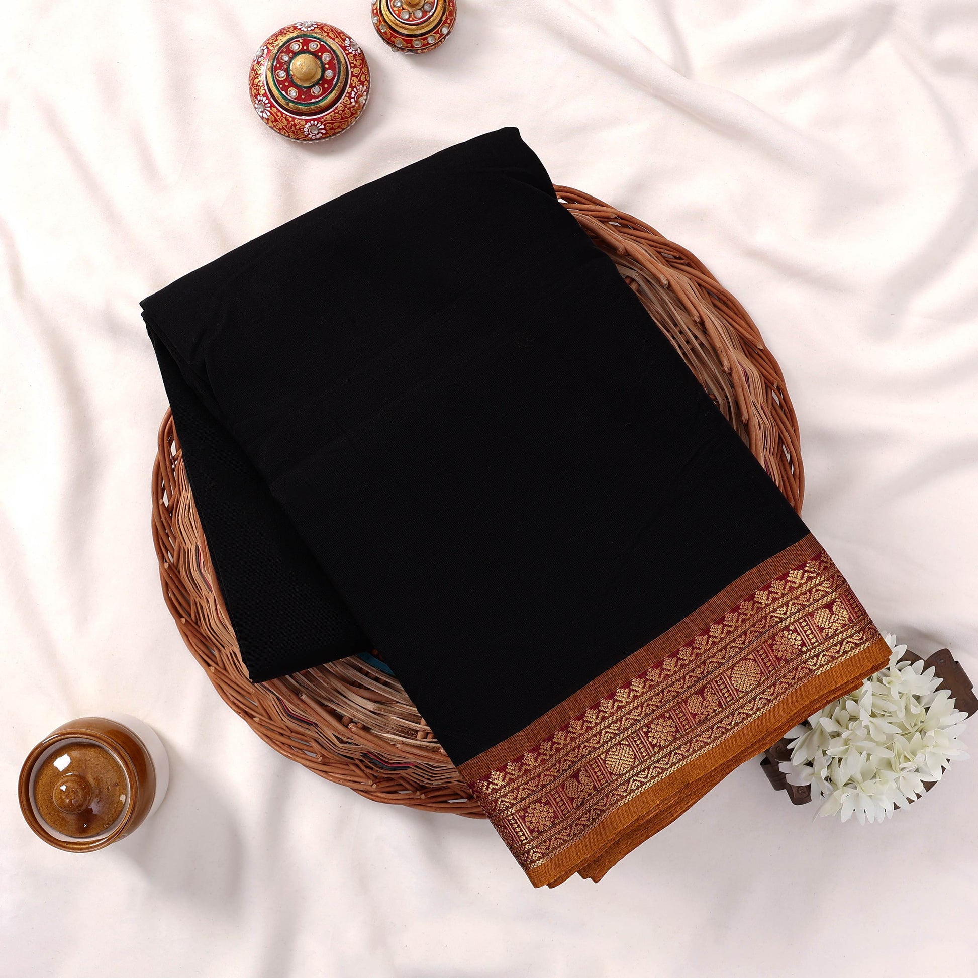 Kanchi Cotton Saree With Black Body And Intricate Gold Zari Border Folded Flat Lay Display In Wicker Basket
