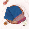 Kanchi Cotton Saree With Blue Body And Contrasting Purple Gold Woven Border Folded Display On Wicker Basket Top View
